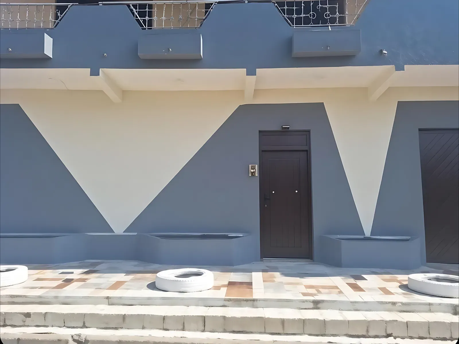 Fachada del local: pared azul grisácea con triángulos crema, puerta oscura en el centro y neumáticos pintados de blanco en la vereda